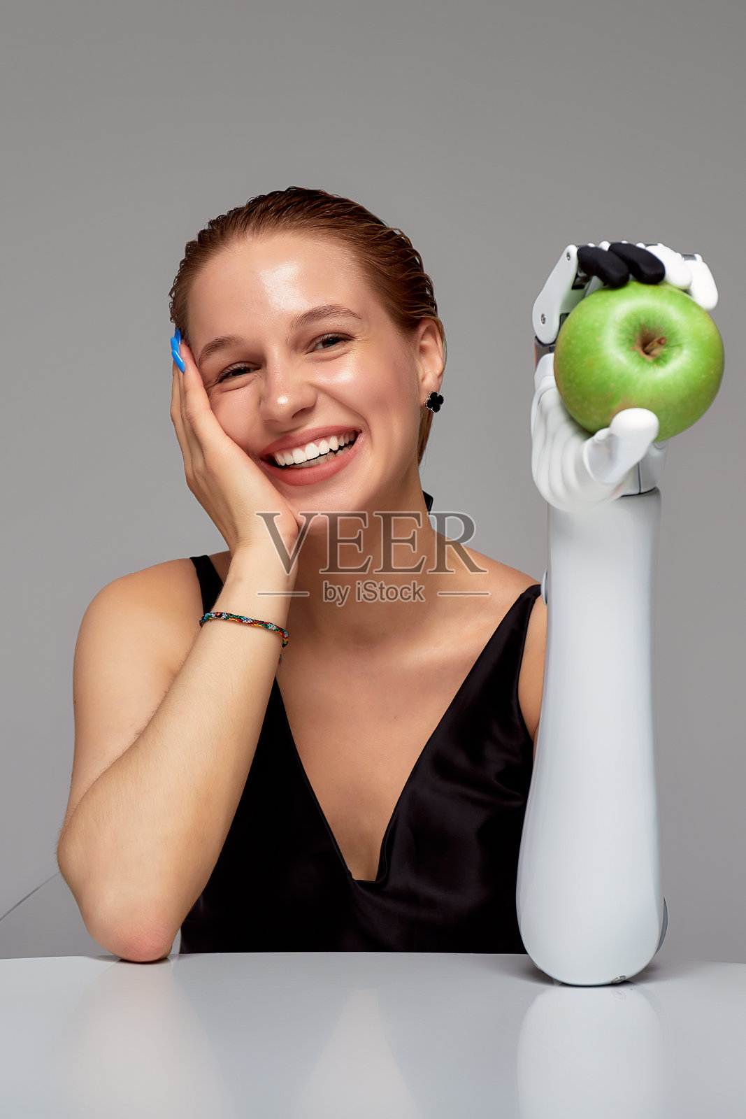 微笑的残疾女孩戴着传感仿生假肢坐在桌子旁。一个美丽的女人用她抬起的人工机械手拿着一个青苹果。失去肢体后的完整生活。照片摄影图片