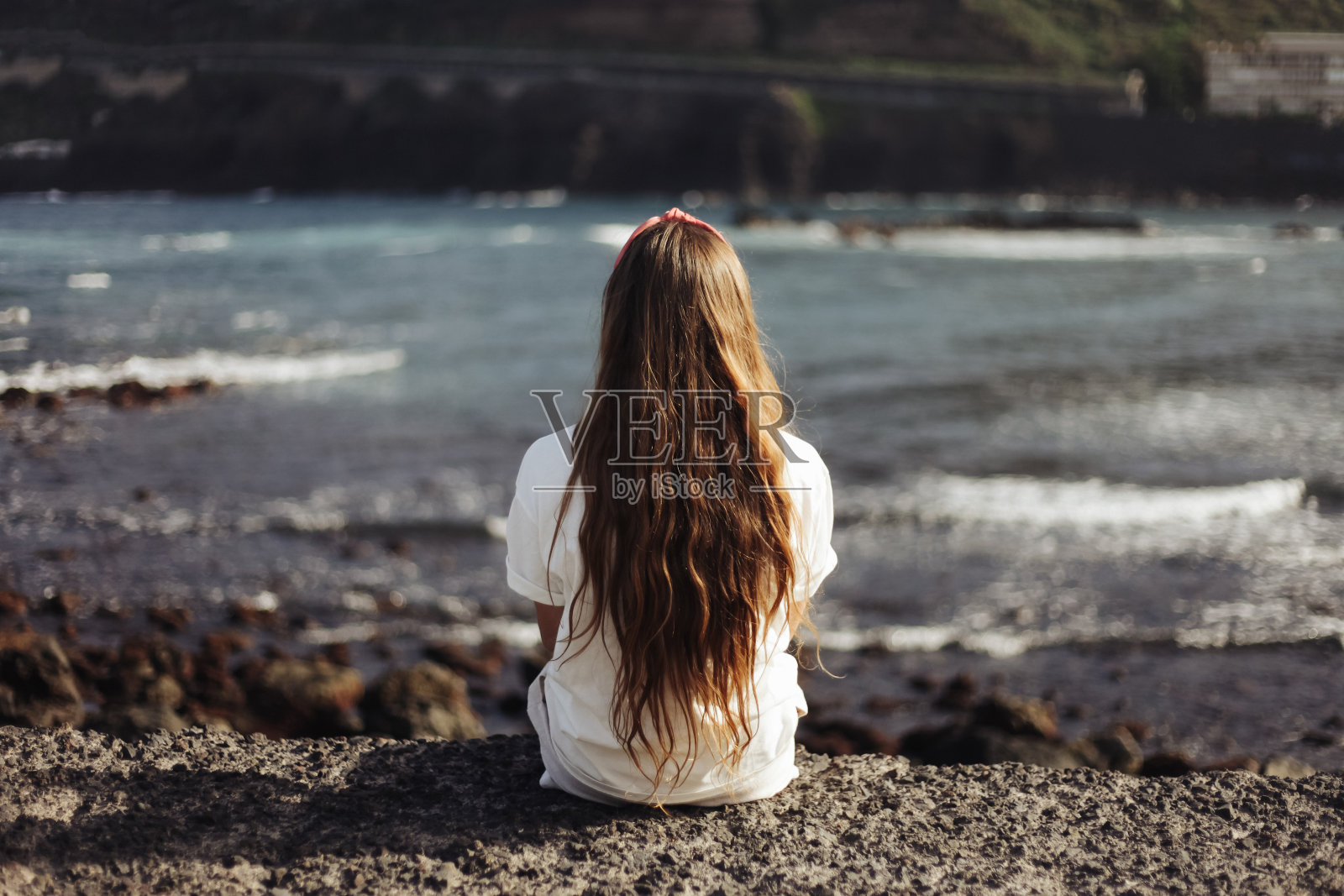 长头发的女人看着波浪。坐在海边。特内里费火山海滩。加那利岛的日落。女孩坐在悬崖边上。照片摄影图片