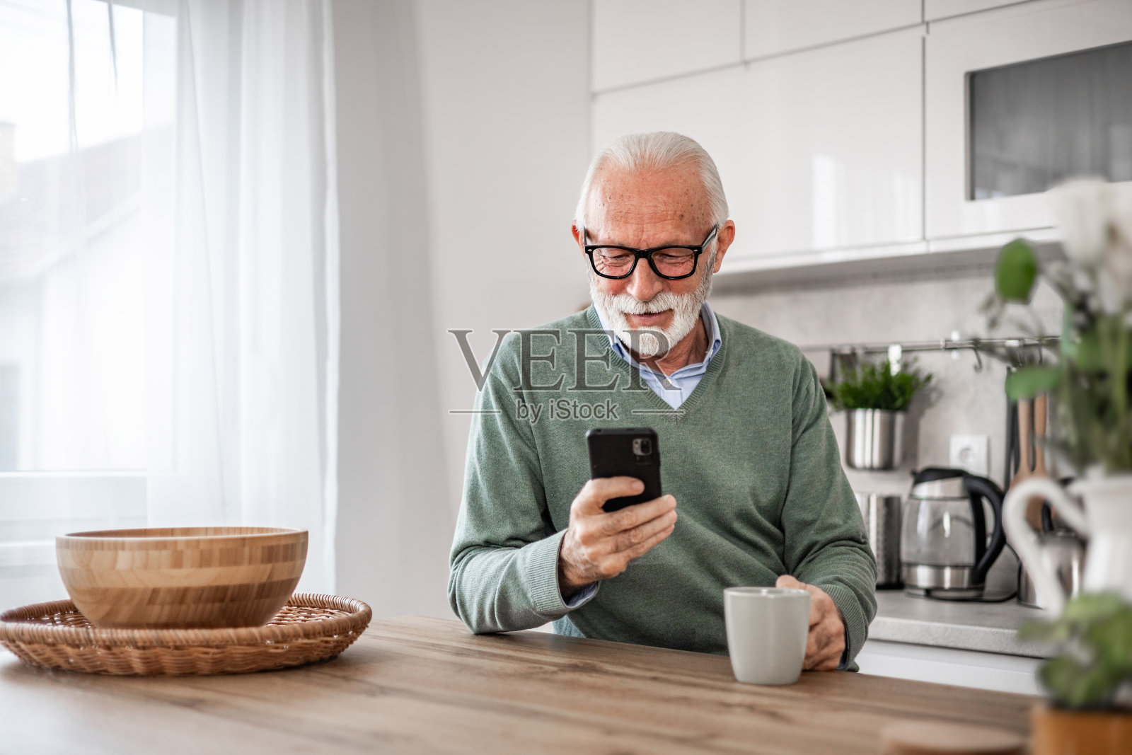 老人,退休老人,坐在厨房里,带着快乐的微笑,喝着热咖啡或茶,用着手机,早晨的例行公事,无忧无虑的退休日子照片摄影图片