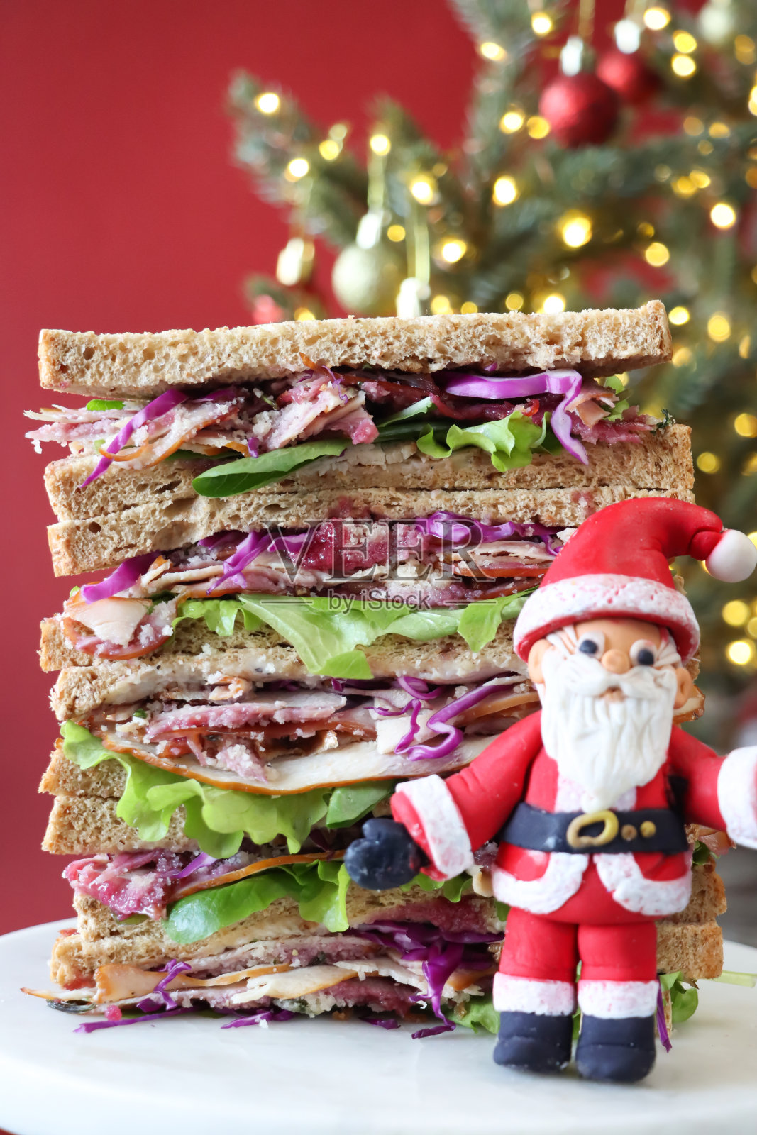 圣诞火鸡三明治的特写形象,展示了馅料,火鸡胸肉片在生菜床上,上面有蔓越莓酱和红卷心菜丝,大理石蛋糕架,装饰的云杉树照片摄影图片