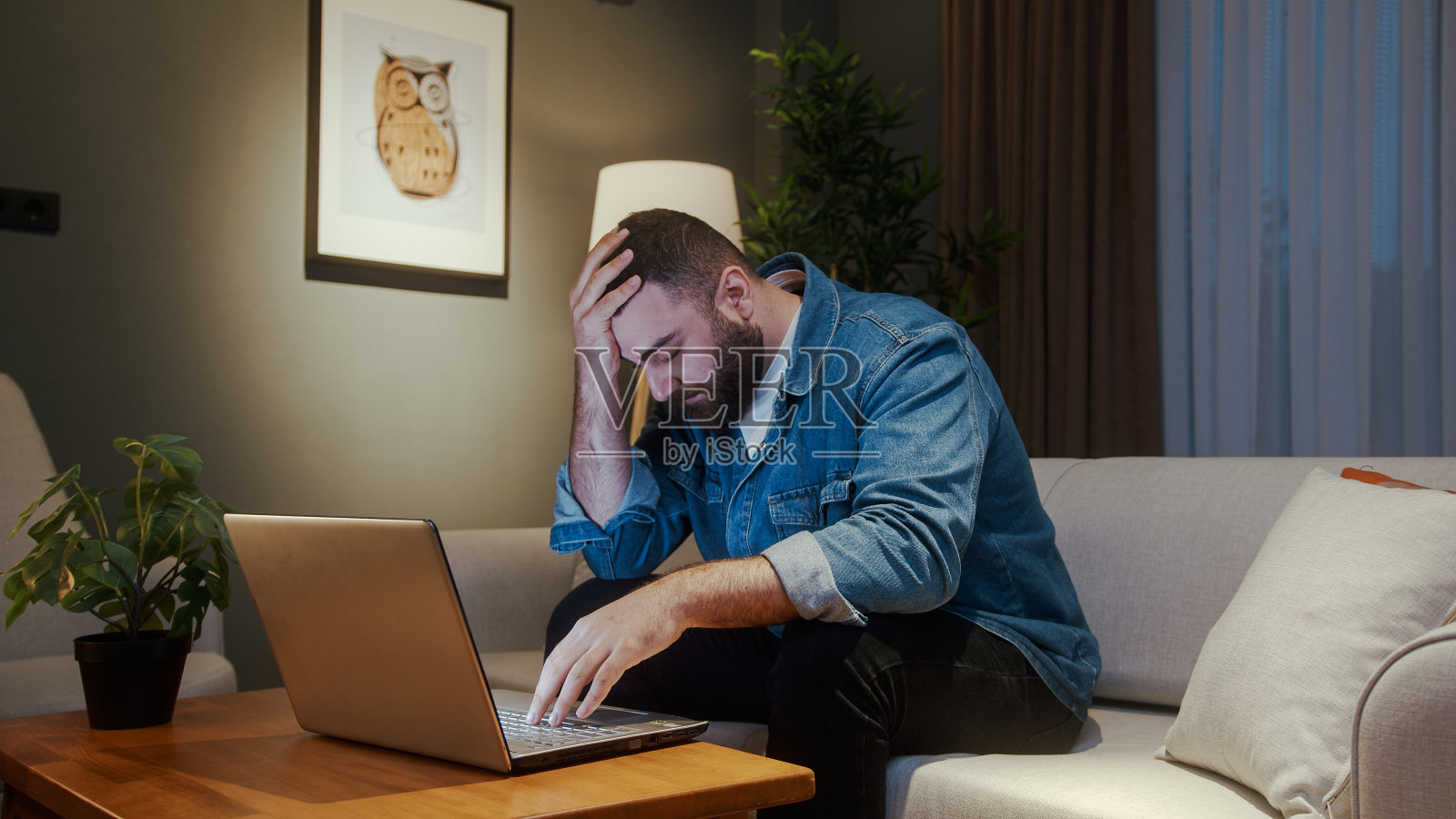 年轻人坐在家里的沙发上,一边使用笔记本电脑,一边感到焦虑和压力。失落的年轻人,电脑里有坏消息照片摄影图片