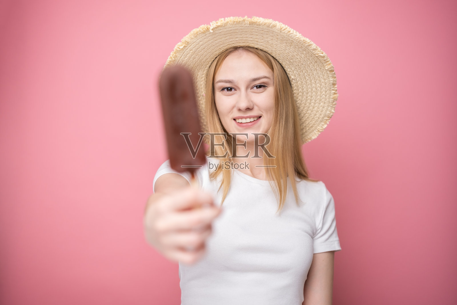 快乐的年轻女子戴着草帽,站在粉红色的背景上吃冰棒。夏日、度假、热天冷餐的概念。照片摄影图片