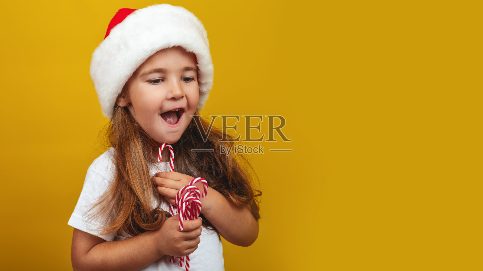 快乐的小女孩戴着圣诞帽吃着圣诞甜筒糖,孤立在黄色背景的工作室里照片摄影图片
