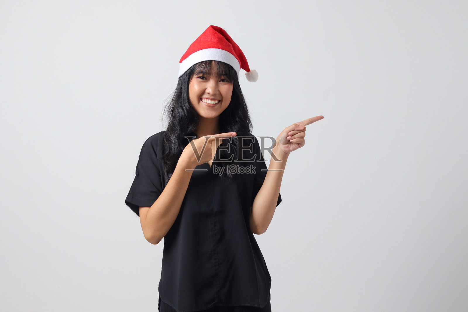有魅力的亚洲女人的肖像,戴着红色的圣诞帽,感觉快乐,用手指指着并展示产品。新年和圣诞节的概念。白色背景上的孤立图像照片摄影图片