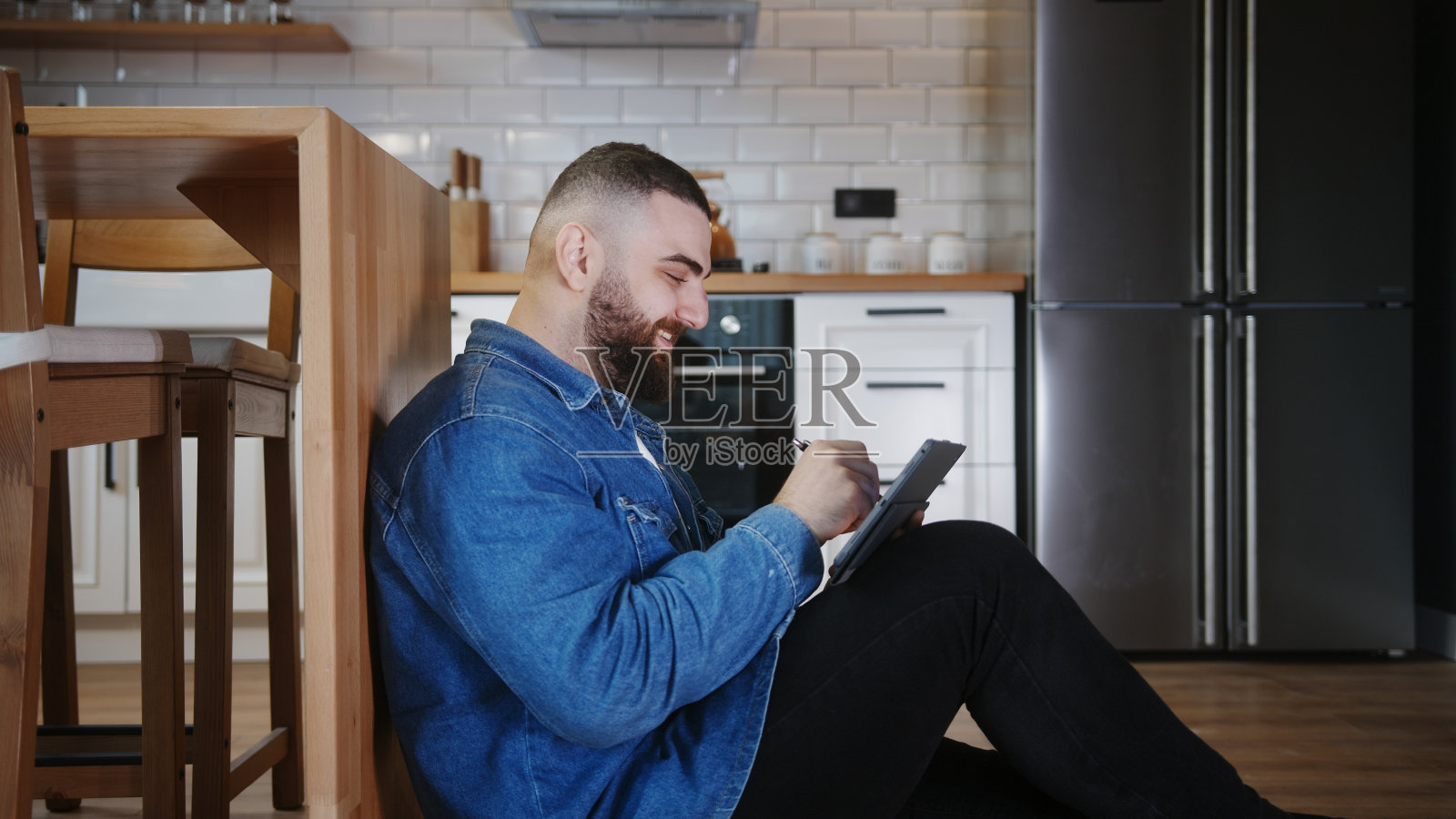 自信的年轻成年商人坐在地板上,靠在厨房柜台上,用平板笔在平板设备的触摸屏上工作和书写照片摄影图片
