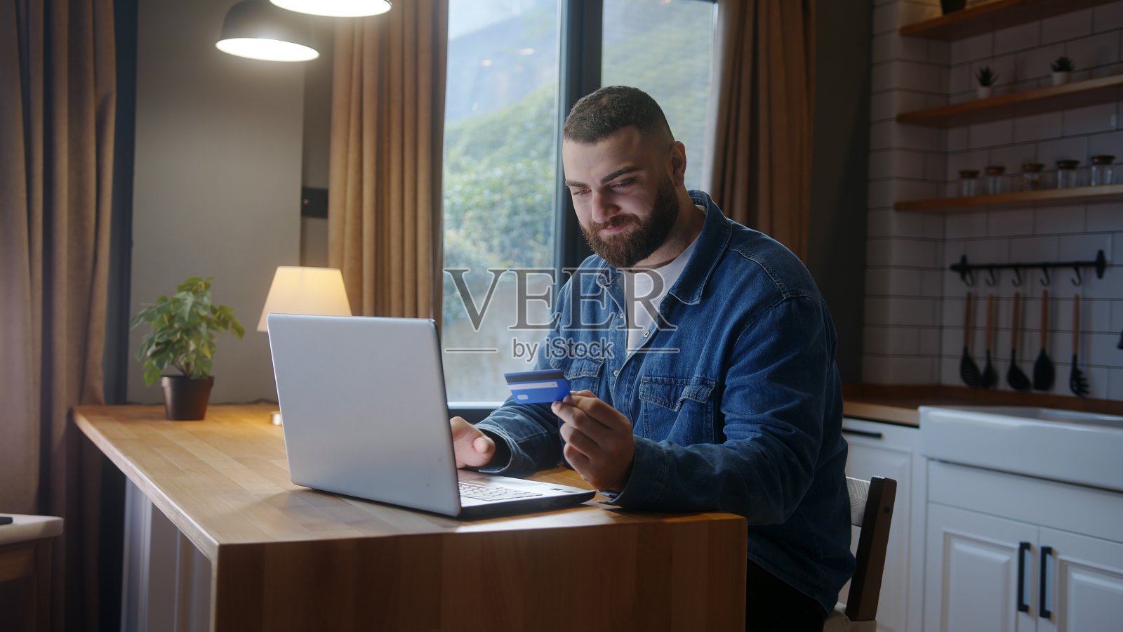 微笑着的年轻男子坐在厨房柜台前,在笔记本电脑上输入信用卡号码,以便在家安全方便地进行远程电子支付照片摄影图片