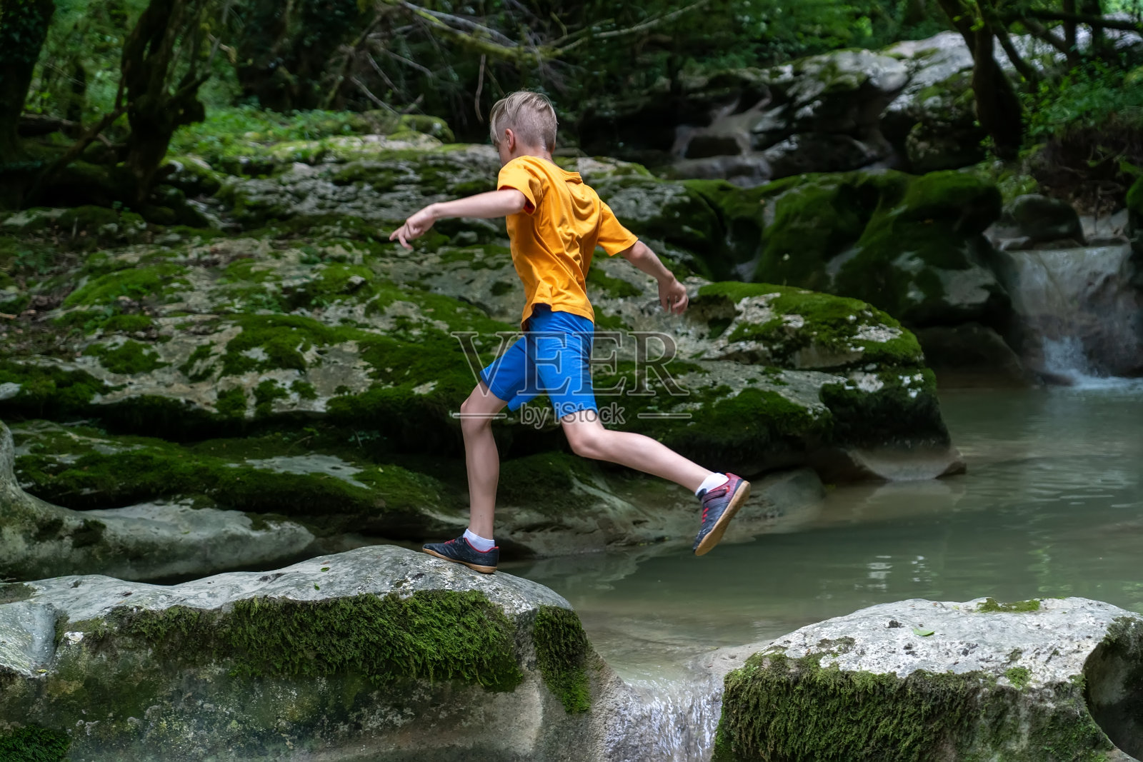 一个活泼的男孩在隐藏在茂密森林中的平静的山河上从一块岩石跳到另一块岩石照片摄影图片