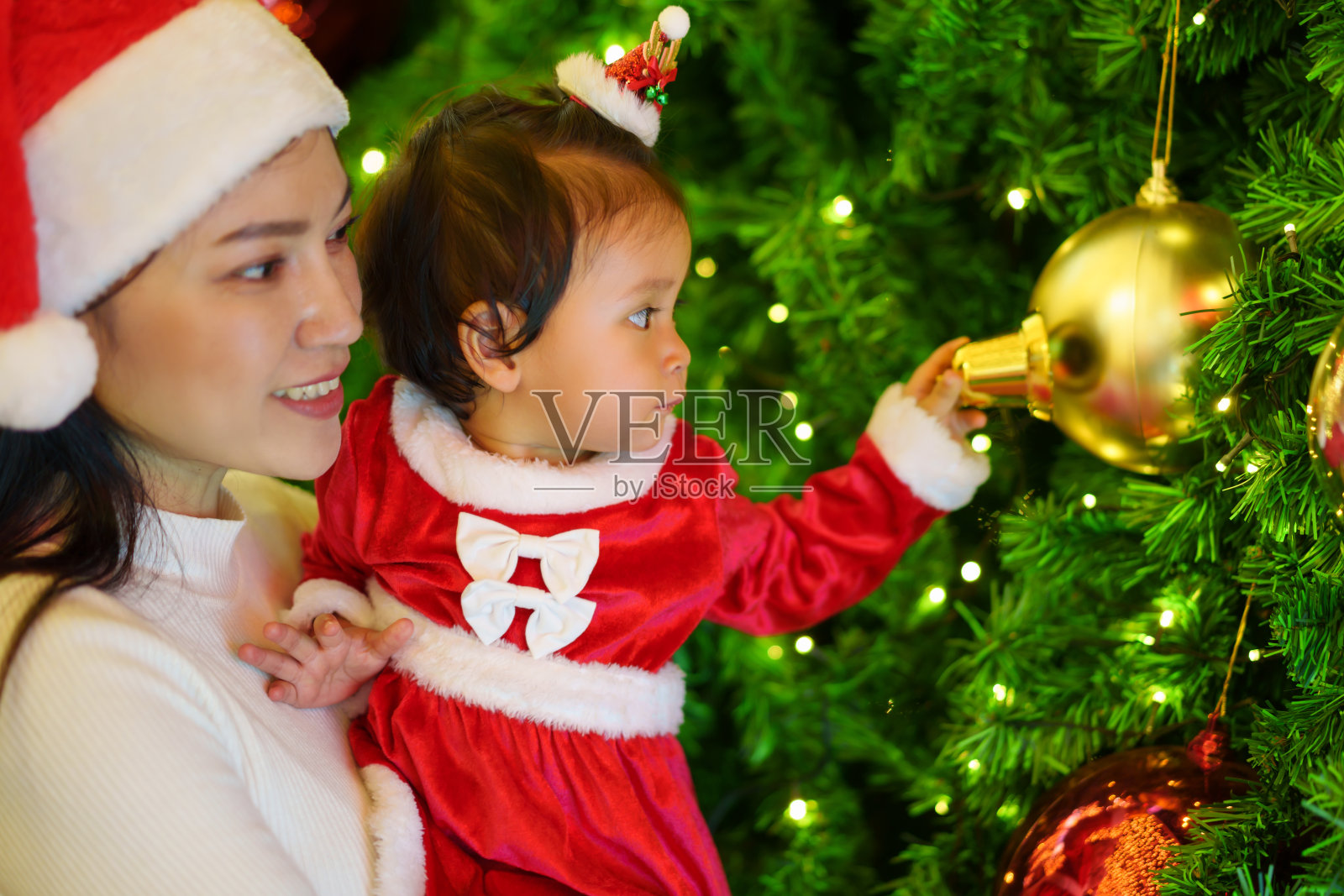 快乐的妈妈和穿着圣诞老人服装的女儿一起玩圣诞球和圣诞树。除夕快乐照片摄影图片