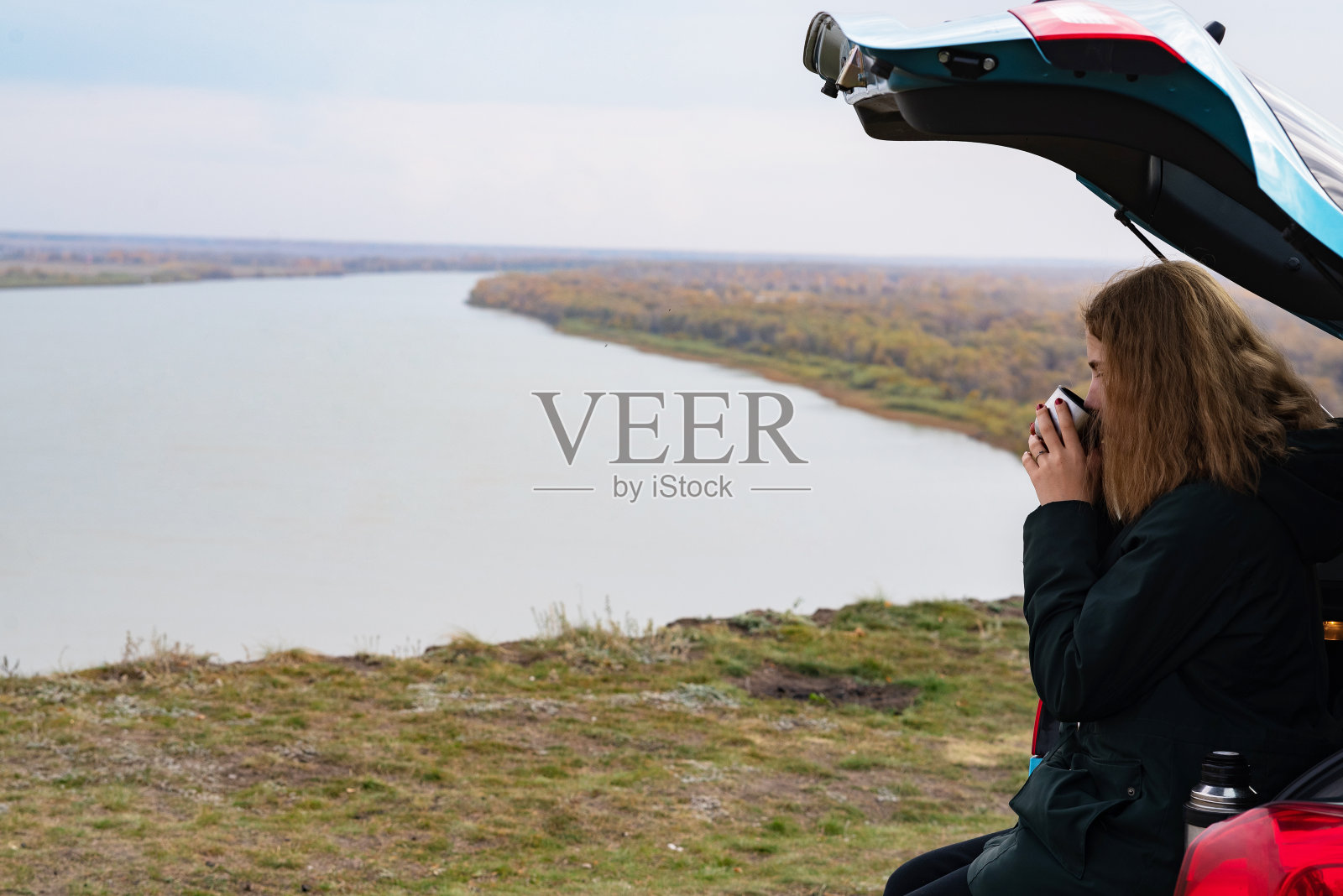 一个穿着毛衣的快乐的年轻白人妇女坐在河边高岸边的汽车后备箱里喝茶照片摄影图片