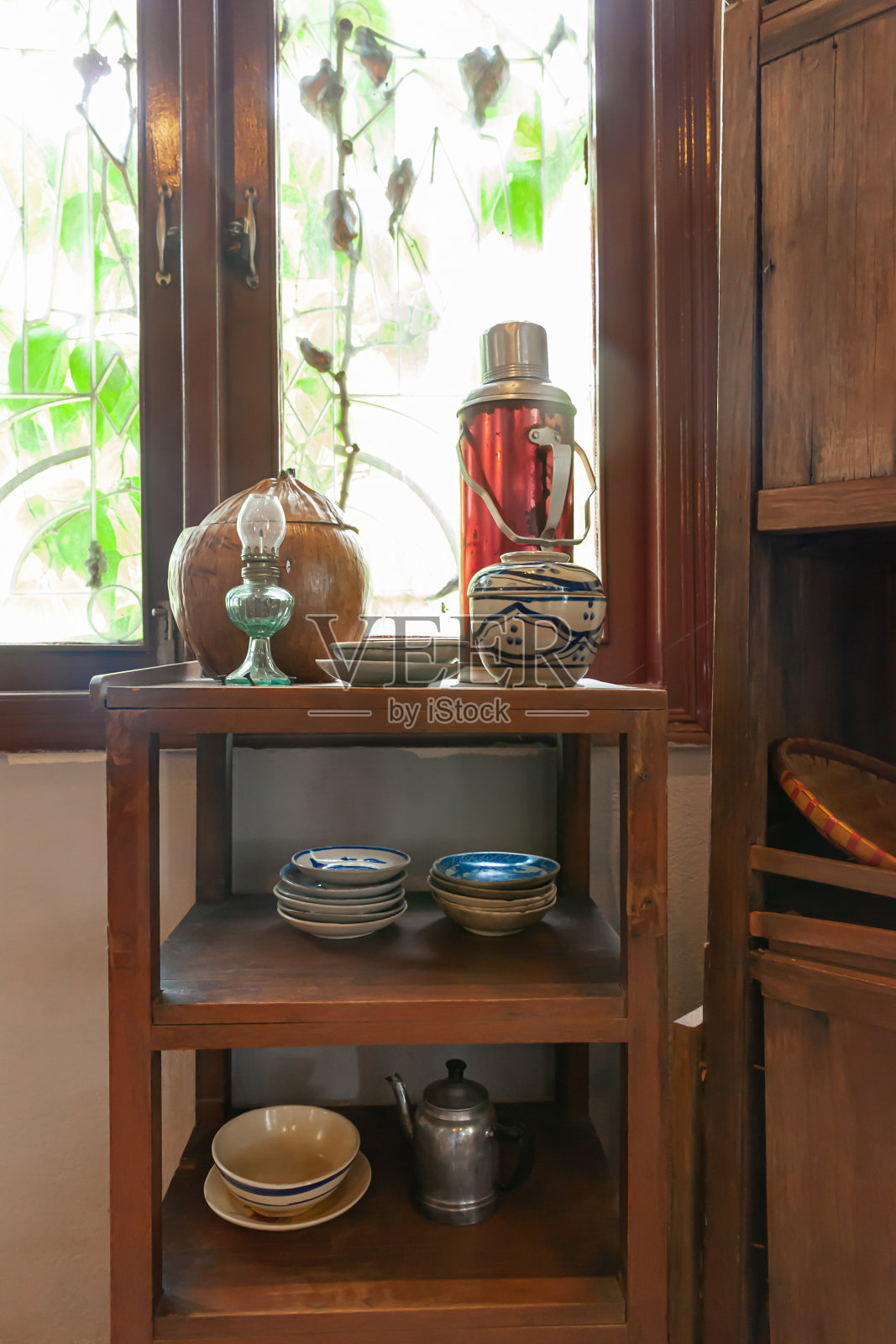 越南厨房内部,瓷花瓶,铝水壶,蓝瓷盘,藤三脚架,油灯,老式保温瓶在一个旧木架子上照片摄影图片