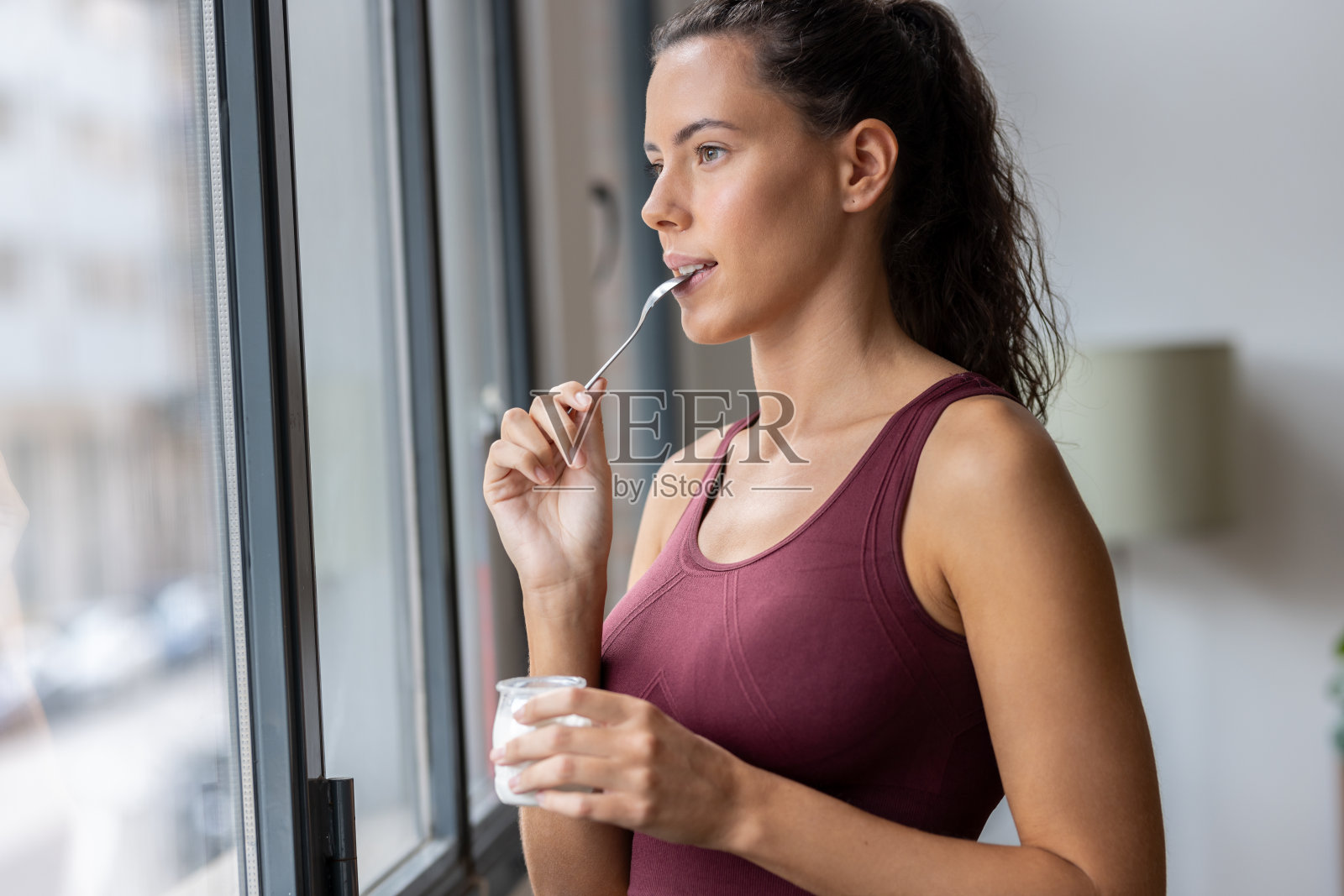 漂亮的运动女人一边吃酸奶,一边在家里透过窗户向前看。照片摄影图片