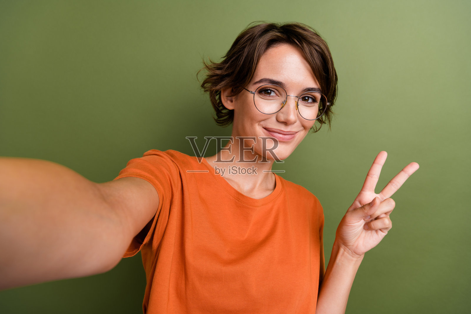 年轻友好的自拍照片,偷拍的女人戴着时髦的眼镜,在卡其色的背景下向订阅者展示v字手势照片摄影图片