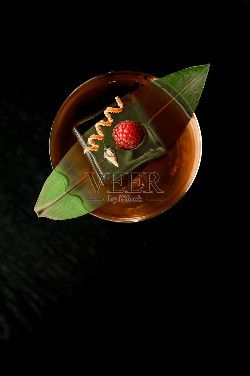 在深色背景上点缀香蕉叶、冰块、覆盆子和柑橘皮的棕色鸡尾酒照片摄影图片