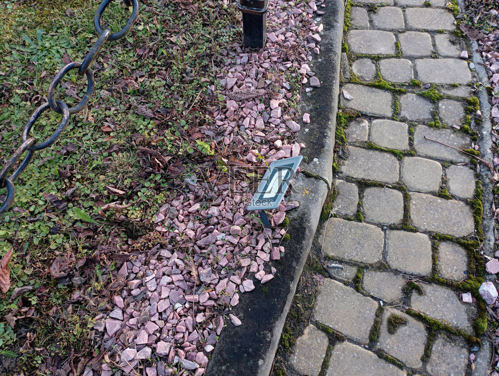 公园狭窄的小路上铺着鹅卵石,两边铺满了砾石。路边有一盏方形的公园灯。附近有一个铁链围栏。照片摄影图片