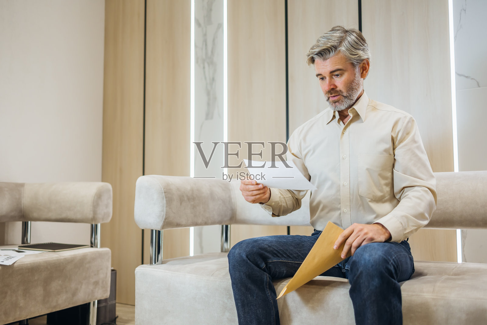 失望的年轻帅哥坐在家里的沙发上读着带坏消息的信。一名穿着衬衫、满脸胡须的男子因收到不愉快的通知而感到沮丧,并被文件中的信息所震惊照片摄影图片