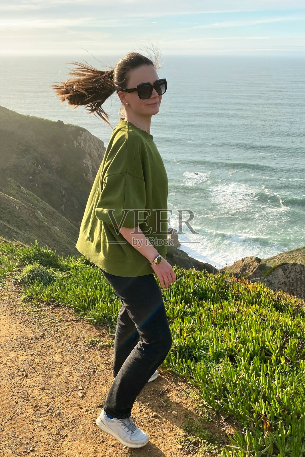 穿着绿色t恤,戴着墨镜的年轻女子站在海边的悬崖上照片摄影图片