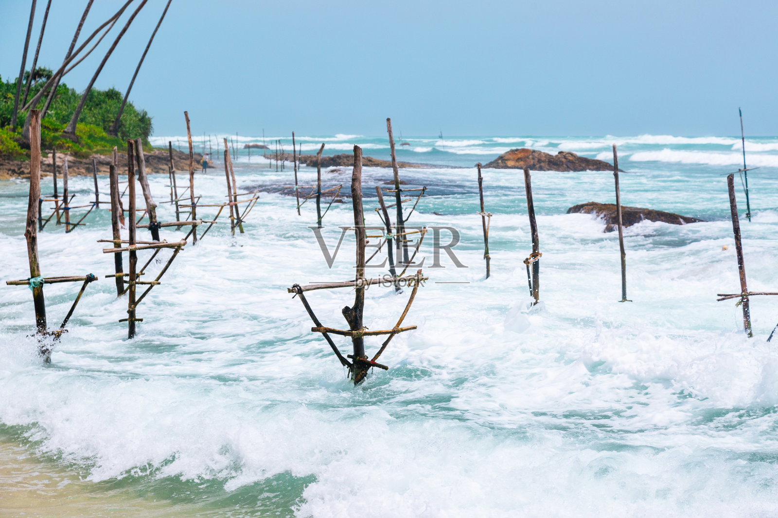 在汹涌的海浪中,斯里兰卡的高跷捕鱼结构,露出岩石的海岸景观和郁郁葱葱的绿色植物。照片摄影图片