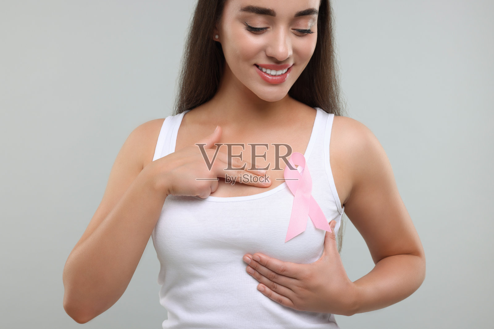 美丽的年轻女子在浅灰色的背景上系着粉红色的丝带。乳腺癌意识照片摄影图片
