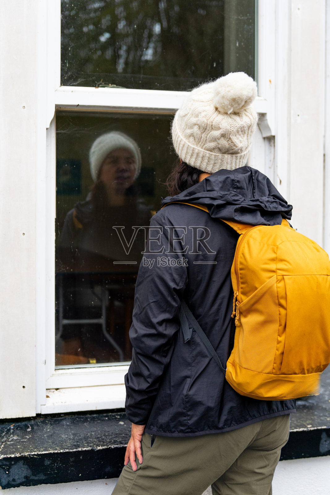 背着双肩包、穿着雨衣的女人透过窗户看着镜子里的自己照片摄影图片