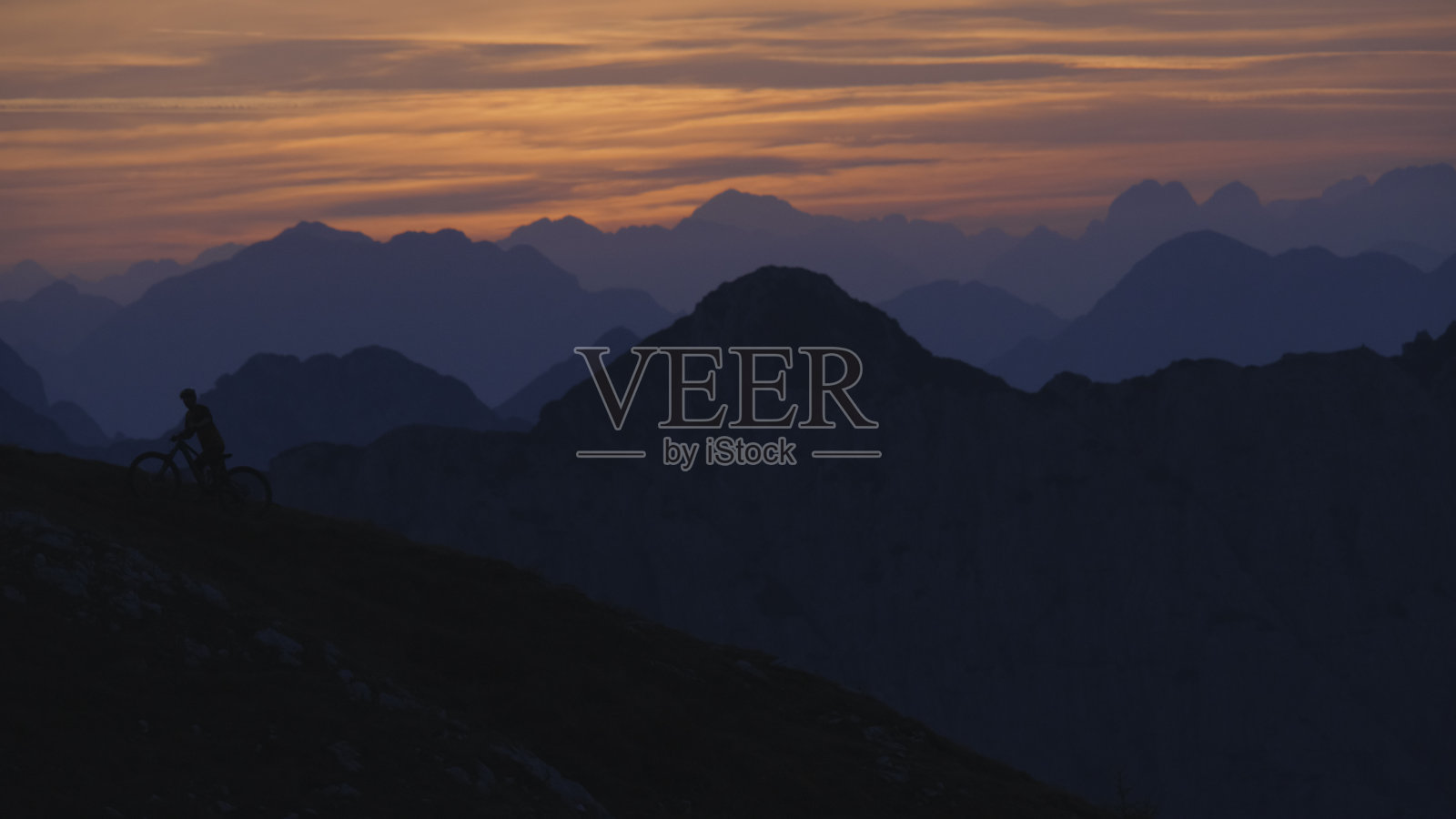 剪影骑自行车的人推自行车,而移动上山对山脉和戏剧性的天空在日落照片摄影图片