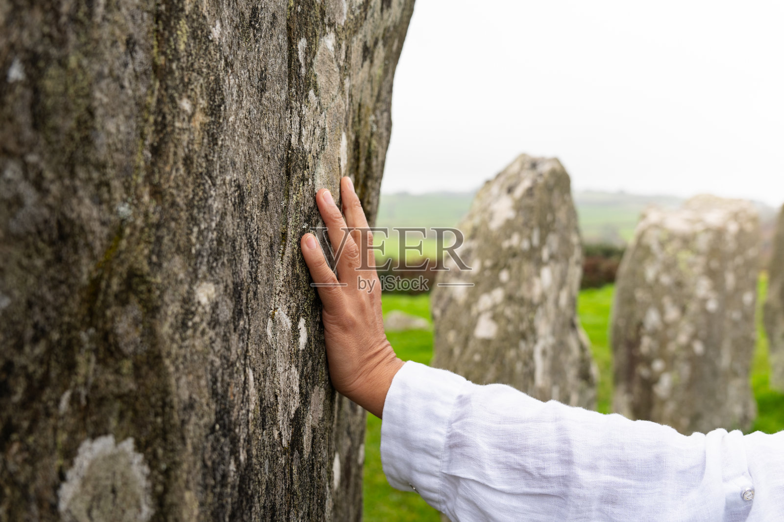 一个女人的手触摸一块巨石的特写,这块巨石属于爱尔兰科克郡的德罗姆贝格新石器时代的圈子,背景是绿色的草地上的其他巨石。照片摄影图片