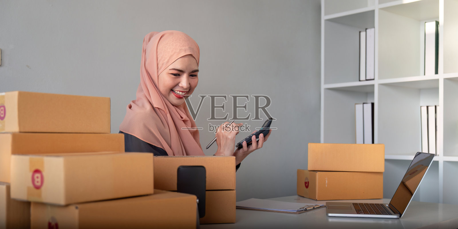 穆斯林妇女在家拿着盒子在网上卖东西。在线销售,接受客户的订单。中小企业经营理念。包裹递送。穆斯林妇女在家使用智能手机和笔记本电脑照片摄影图片