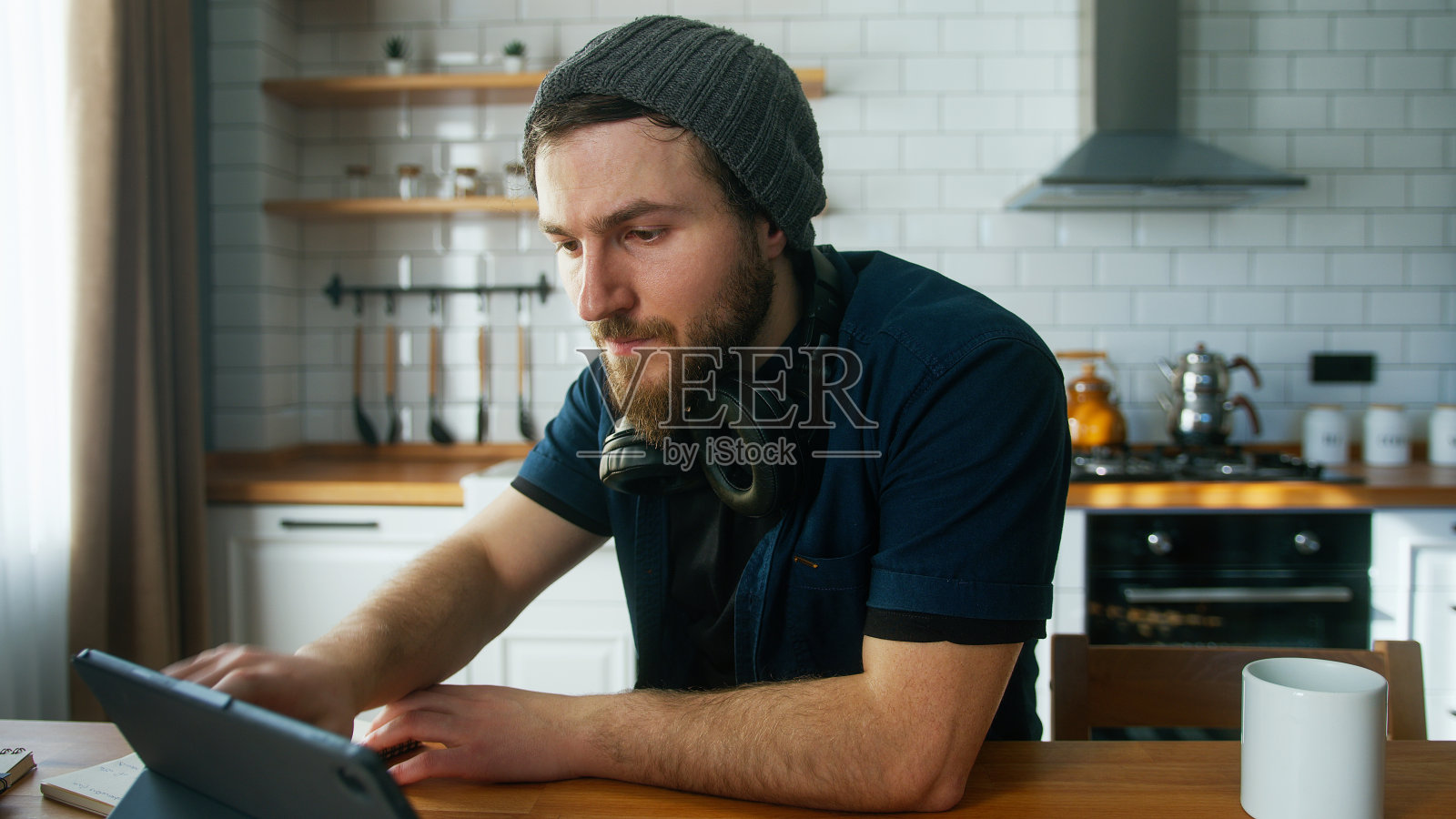 戴着无边帽的商人在家厨房里工作用平板电脑,做笔记,做研究。忙碌的自由职业者在现代科技笔记本设备上工作照片摄影图片
