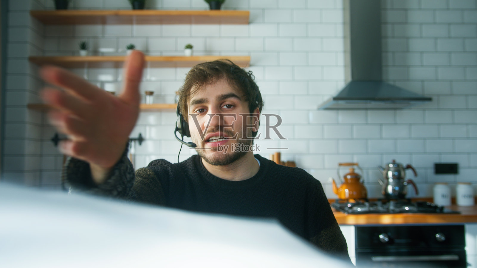 年轻愤怒的经理带着无线耳机坐在家里的现代化厨房里,说话,在视频电话会议中展示报告,对员工生气,扔文件照片摄影图片