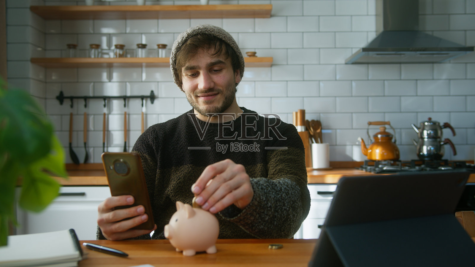 一个面带微笑、头戴无边帽的男人坐在家里的现代厨房里,手里拿着智能手机,看着相机,往扑满里放了几枚硬币照片摄影图片