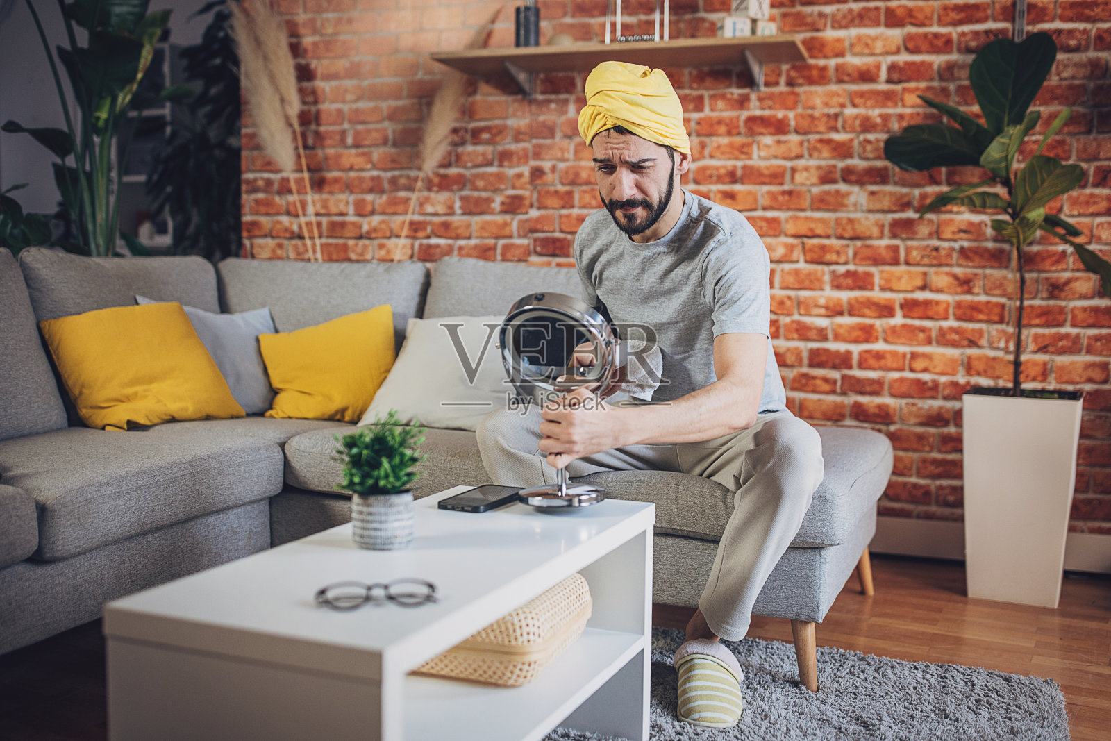 男人洗澡后坐在沙发上,头上裹着毛巾,看着镜子里的痘痘照片摄影图片