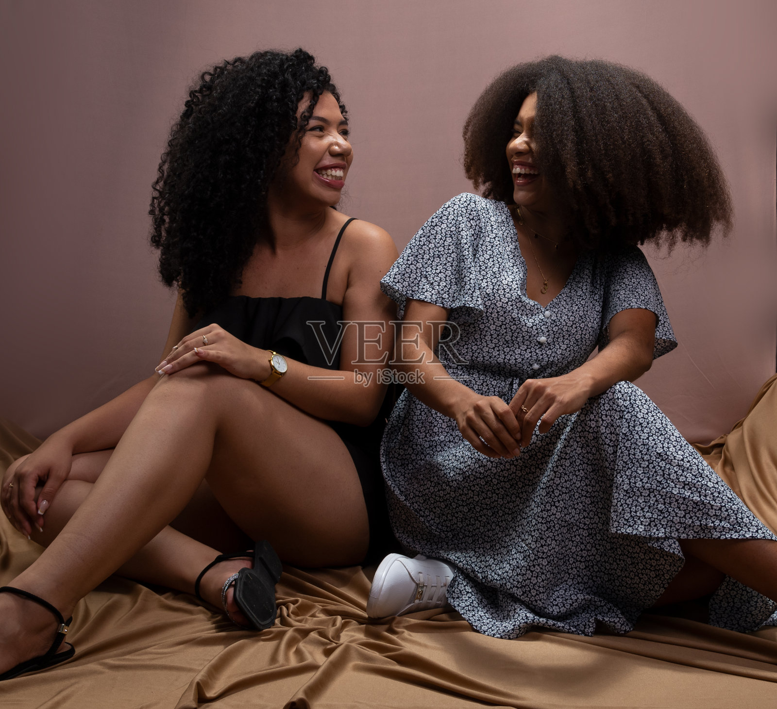 两个年轻,快乐,美丽的朋友,坐在鲑鱼色的背景上,彼此看着对方。照片摄影图片