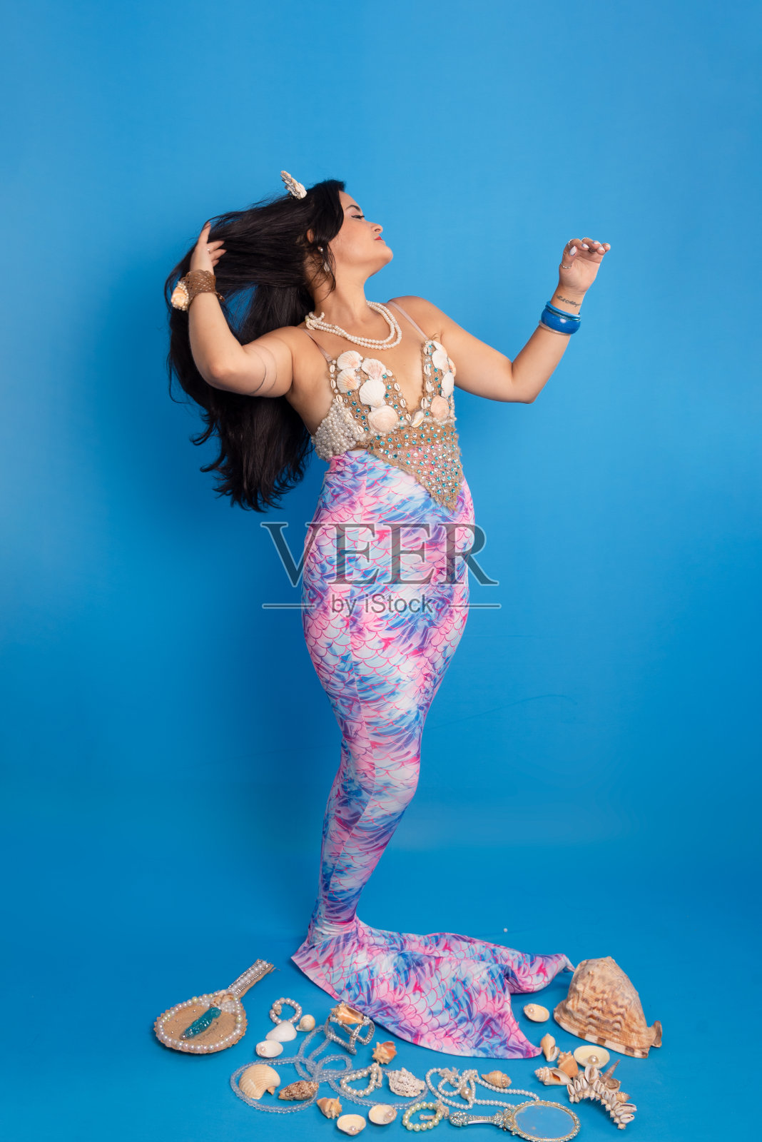 美丽的女人,戴着由贝壳制成的头饰和手镯,在蓝色的背景下,打扮成美人鱼,旁边有几个贝壳,一面镜子和珍珠项链抚摸着她的黑发。照片摄影图片