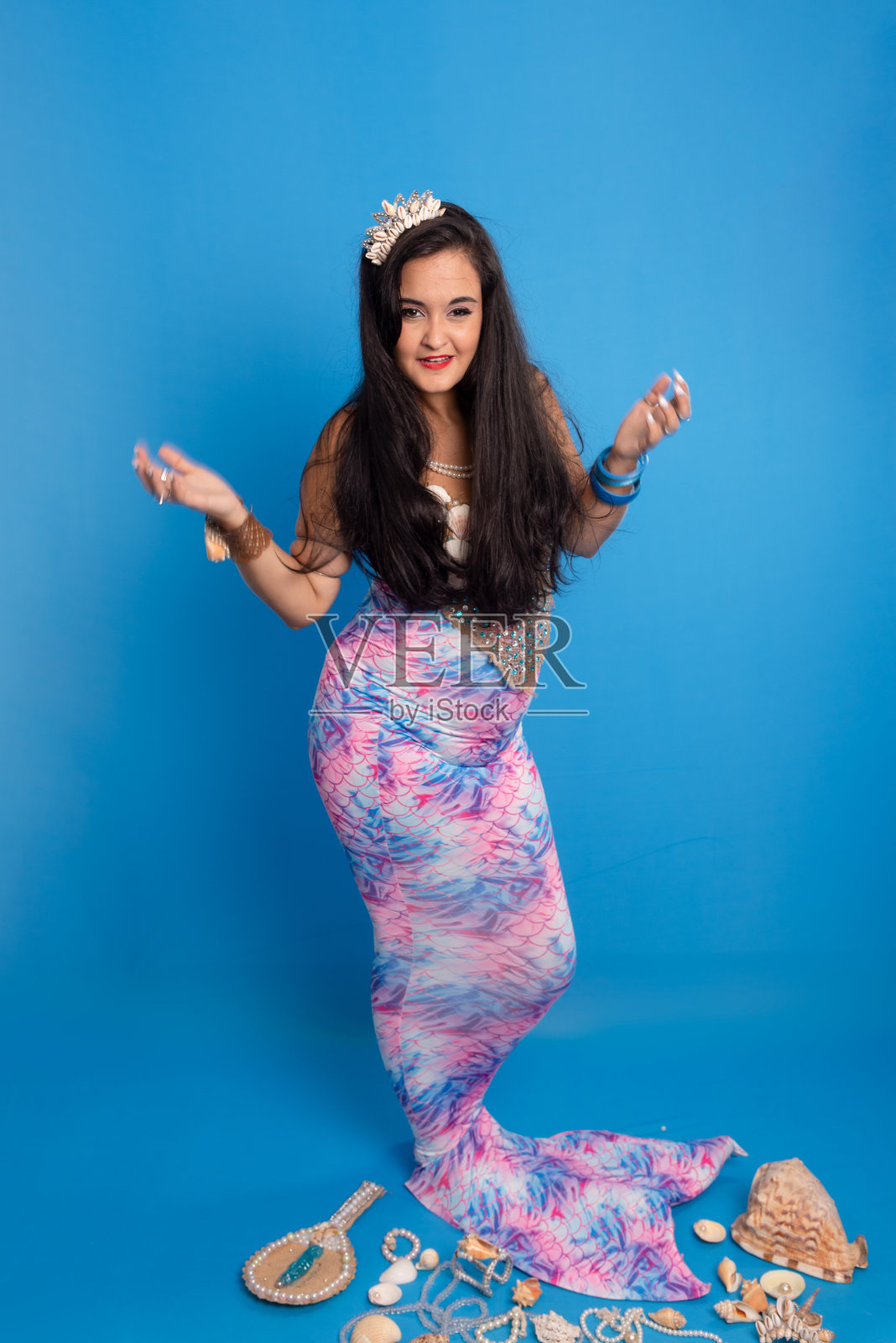 美丽的女人,戴着头饰和海洋饰品,衬着蓝色的背景,旁边有几只贝壳,珍珠项链和一面镜子,打扮成美人鱼。照片摄影图片