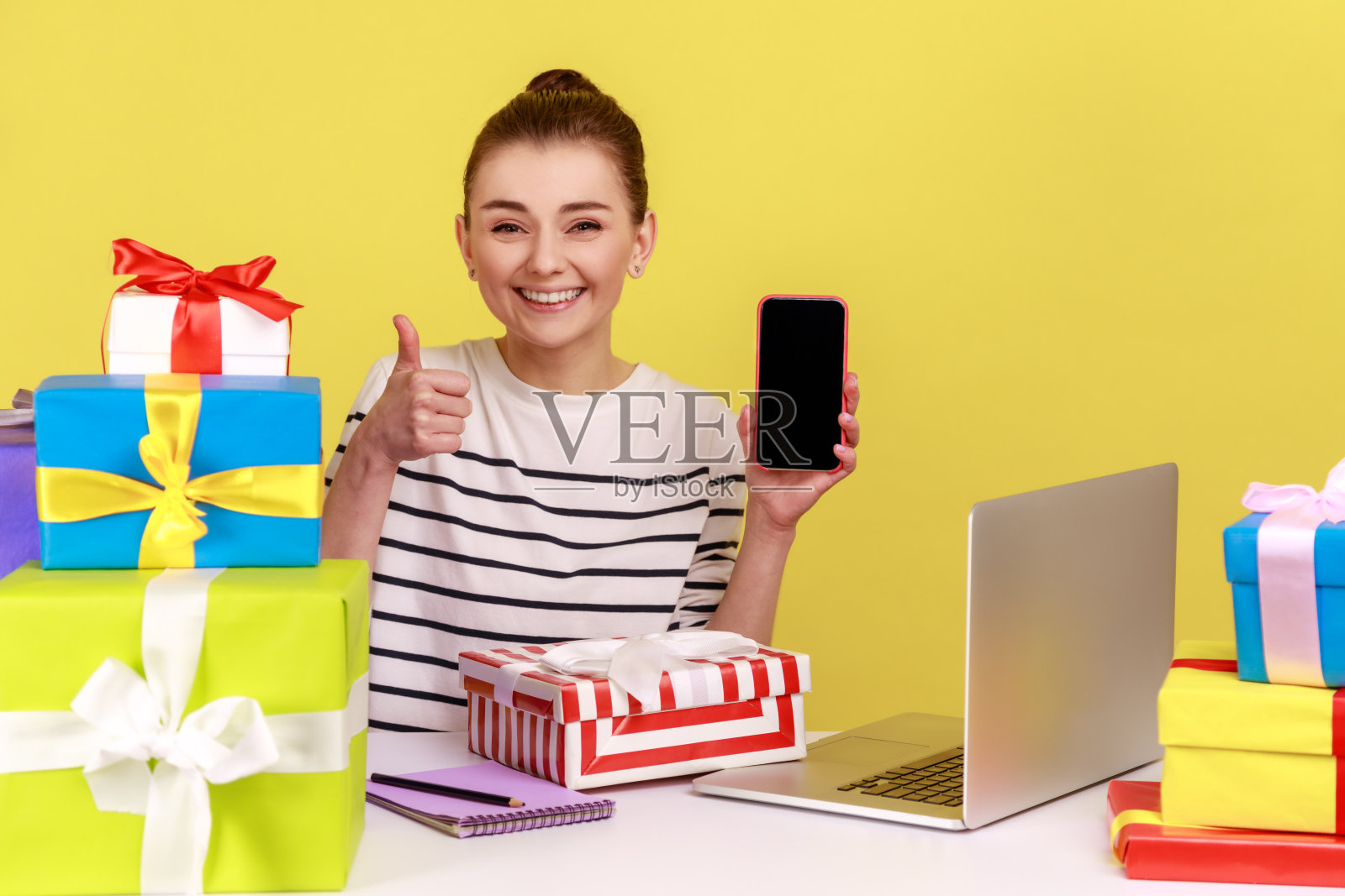 女人拿着礼品盒坐在工作场所,拿着竖起大拇指的空白手机照片摄影图片