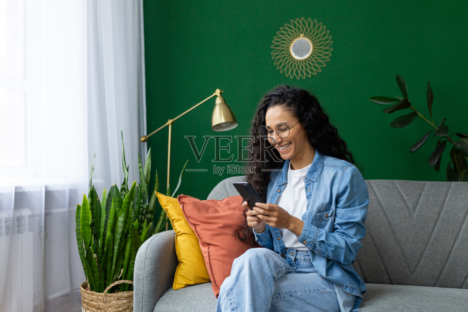 美丽的西班牙裔妇女在客厅的沙发上,面带微笑,手里拿着电话,使用智能手机上的应用程序,浏览互联网和社交网络照片摄影图片