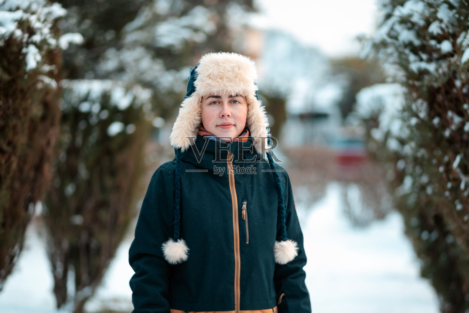 一个年轻的白人妇女的肖像,穿着夹克,戴着带耳盖的帽子,在积雪覆盖的小巷里摆姿势照片摄影图片