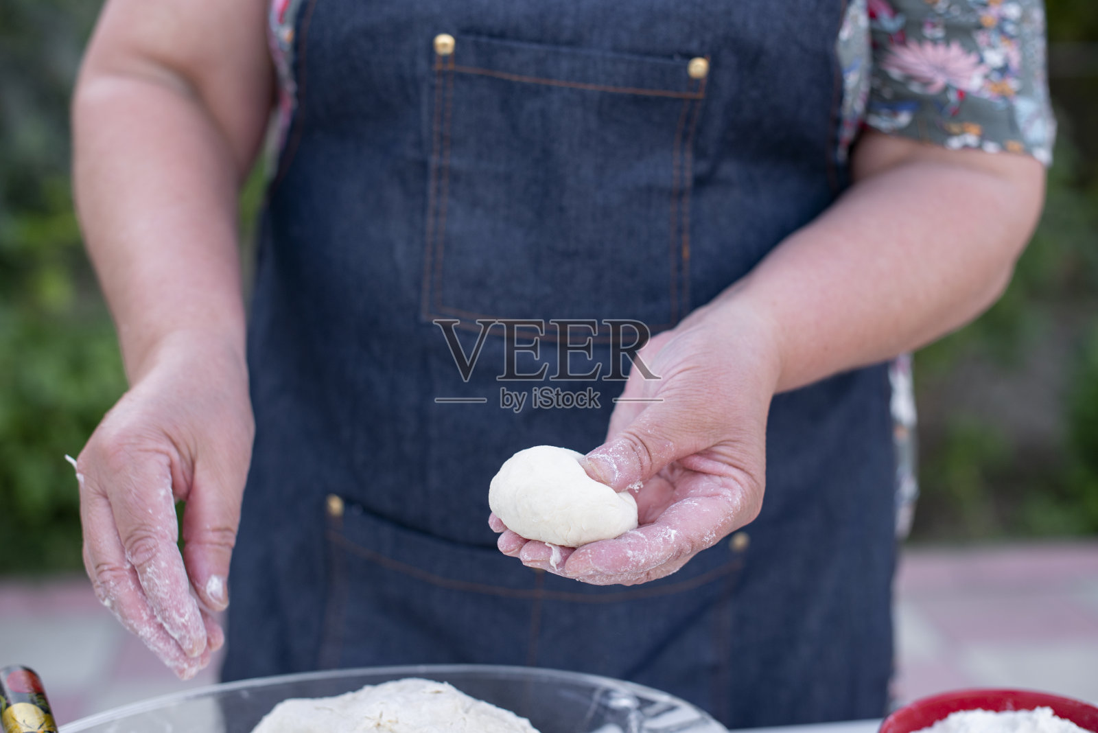 老妇人戴着蓝围裙,在户外揉面、捏面、制作、擀馒头。照片摄影图片