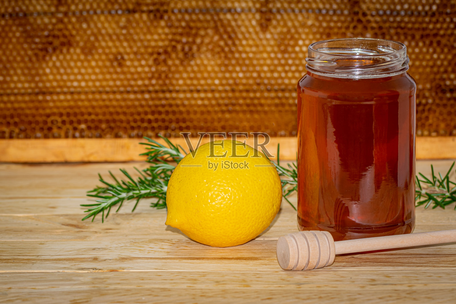 一罐没有盖子的蜂蜜,分配器,迷迭香和一个柠檬放在木头上,背景是蜂巢照片摄影图片