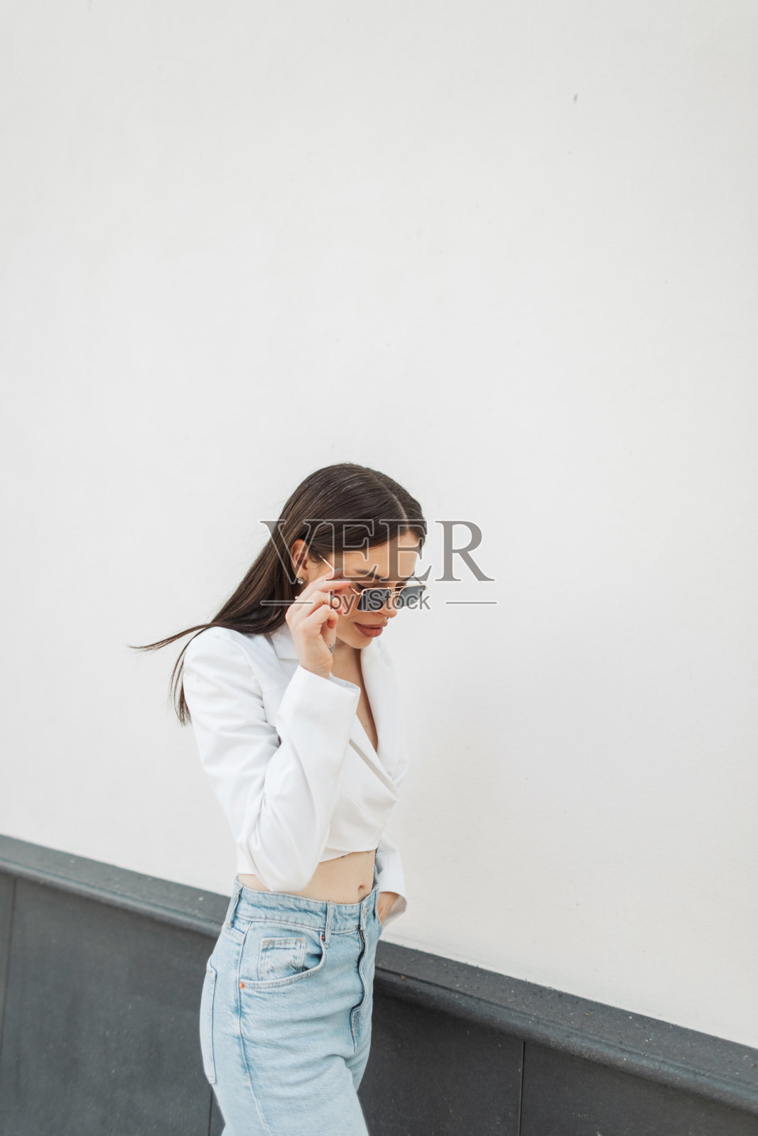 漂亮时髦的女人穿着时尚的衣服,白色的夹克和蓝色的牛仔裤,戴上复古的太阳镜,走在城市的白色建筑附近照片摄影图片