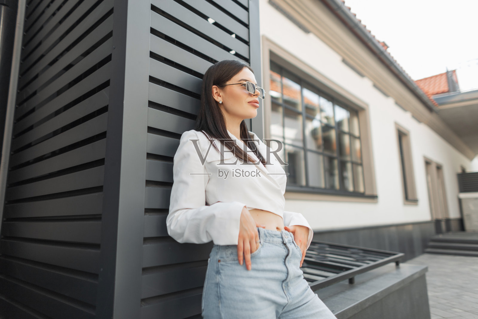 美丽时尚的都市女性肖像,带着复古太阳镜,穿着时髦的外套和高腰牛仔裤,站在街道上的建筑物附近照片摄影图片