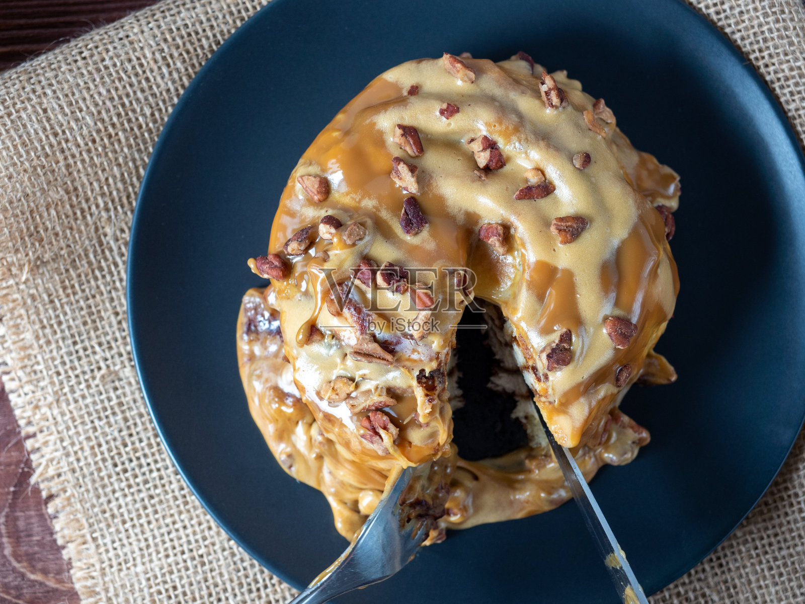 一个特写:一个涂着焦糖和坚果装饰的肉桂面包放在一个黑色盘子和一张亚麻餐巾上,甜点是用刀叉切开的。俯视图,平铺照片摄影图片