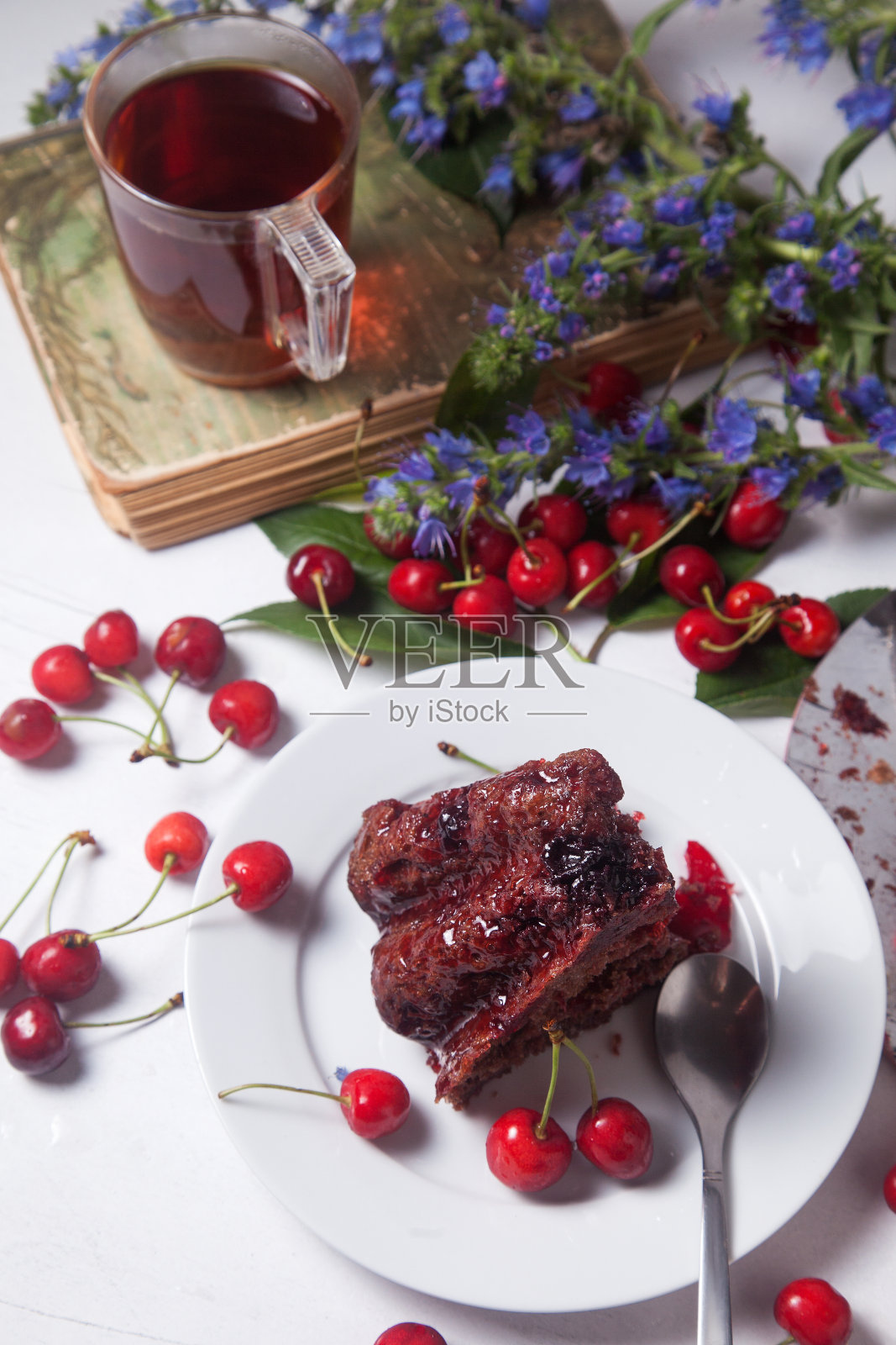 巧克力蛋糕配甜樱桃,古书上的一杯茶,白色背景上的一束蓝花。照片摄影图片