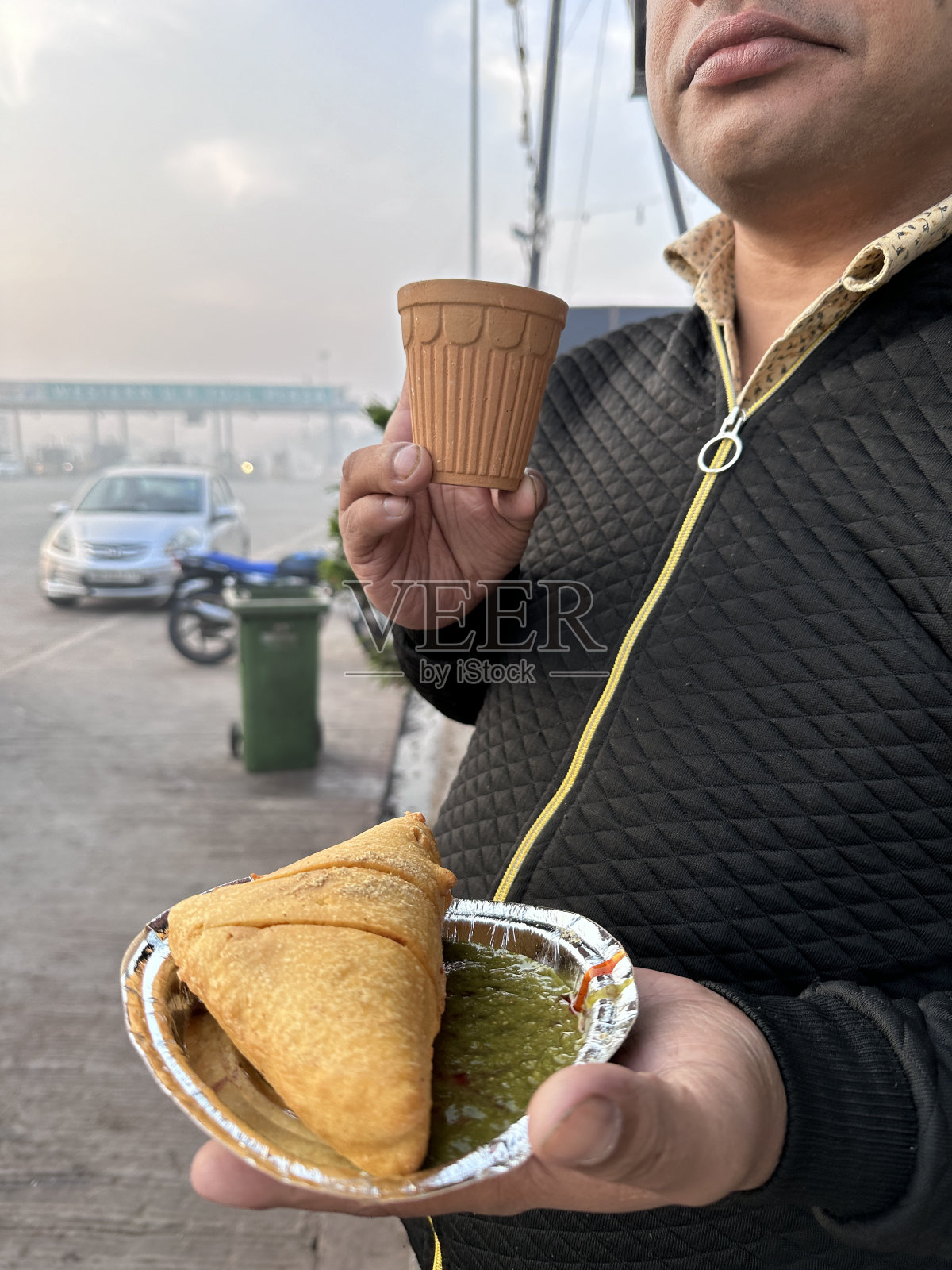 一名无法辨认的男子手持一次性锡纸盘子,里面塞满了五香土豆、豌豆和肉,配上薄荷香菜蘸料和街头食品市场摊位上的马萨拉茶,这是前景的焦点照片摄影图片