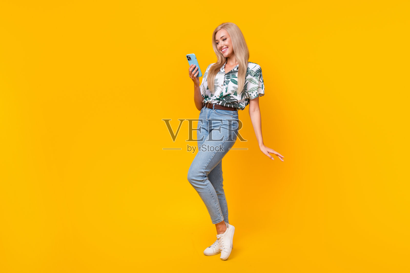 可爱女孩的全身照片穿着时尚的衬衫牛仔裤看智能手机阅读短信孤立的黄色背景。照片摄影图片