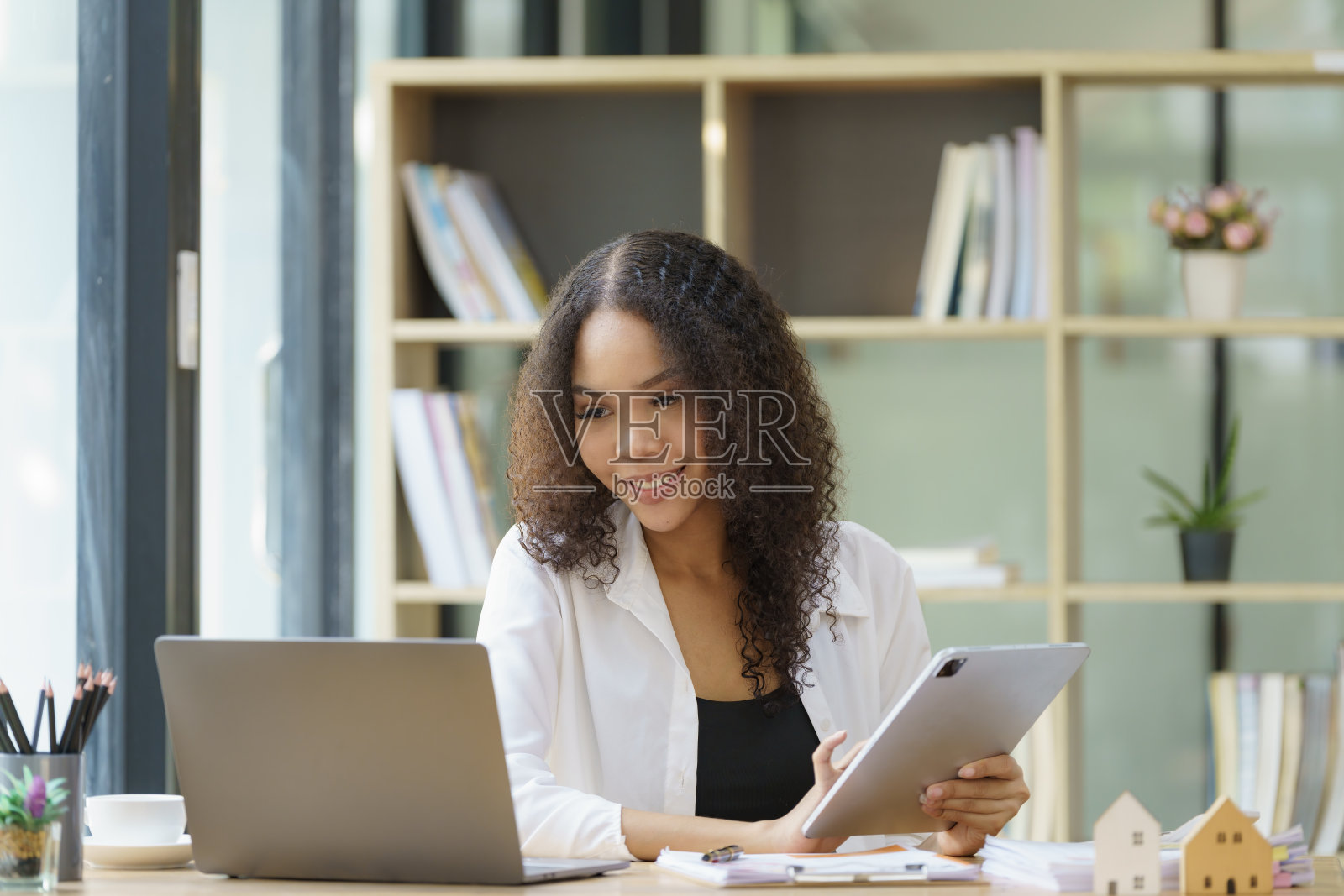 年轻的非洲裔女商人公司经理销售主管使用数字平板电脑工作,坐在办公室的办公桌前看着笔记本电脑思考。照片摄影图片