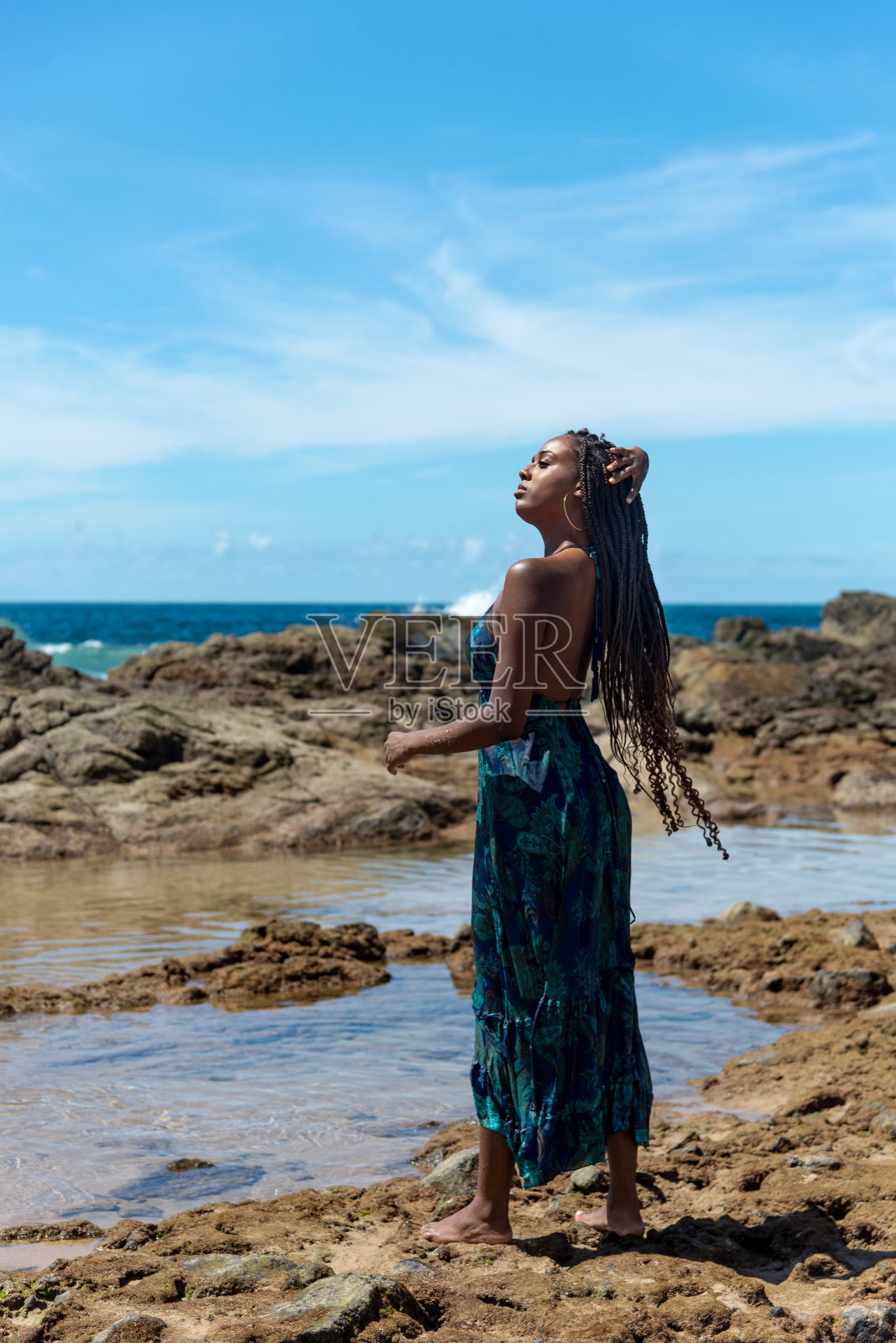 美丽的女人站在沙滩上的岩石上,穿着蓝色的衣服,抚摸着她的辫子。照片摄影图片
