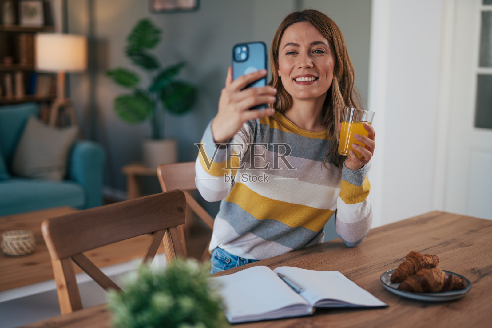 友好的女士在家里用智能手机说话。放松的女人坐在餐厅喝果汁。照片摄影图片