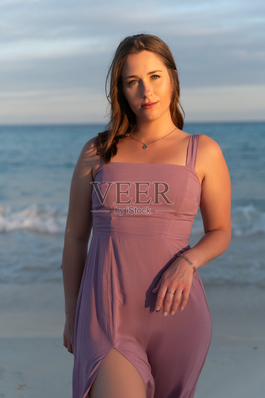 日落时分,身穿粉色连衣裙的女子走在沙滩上。照片摄影图片