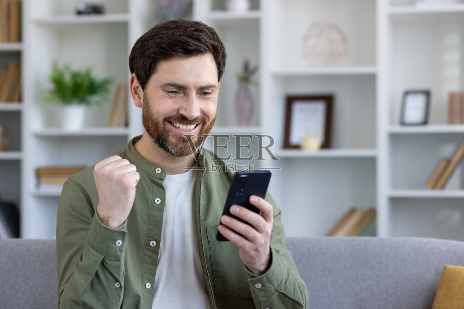 男主在手机上收到在线中奖通知和成功的在线成就结果,坐在家里客厅的沙发上,赢家举起手,做出胜利的手势照片摄影图片