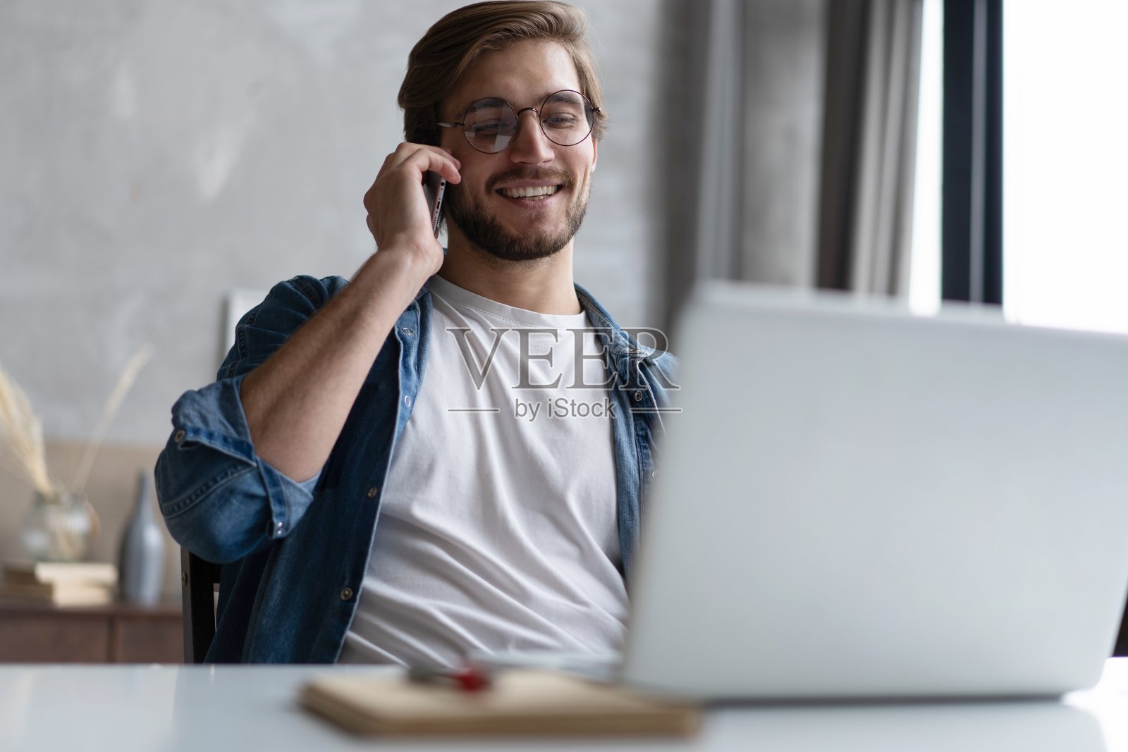 开朗的年轻人在家办公,用手机和笔记本电脑远程工作,坐在房间的办公桌前照片摄影图片